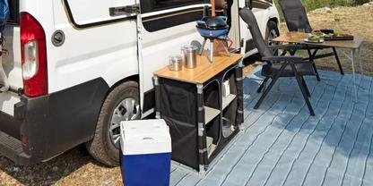 Wei&szlig;er Campervan neben klappbarem K&uuml;chenregal mit Holzarbeitsplatte und drei Edelstahlbechern, blau-wei&szlig;e K&uuml;hlbox auf dem Boden, hellblauer Bodenbelag, zwei Klappst&uuml;hle und kleiner Beistelltisch