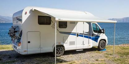 Wei&szlig;es Wohnmobil mit ausgefahrener Markise auf kiesigem Ufer neben dem Meer, Berge im Hintergrund