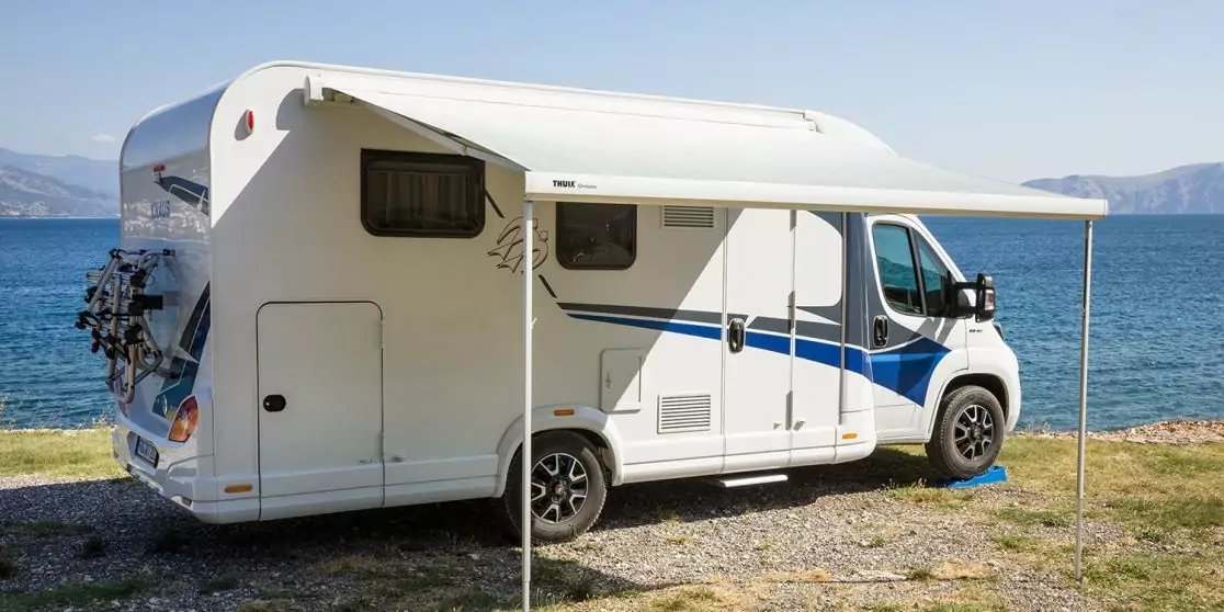 Wei&szlig;es Wohnmobil mit ausgefahrener Markise auf kiesigem Ufer neben dem Meer, Berge im Hintergrund