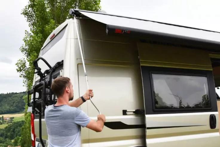 Mann in grauem T-Shirt spannt eine Markise an der Seite eines Wohnmobils; Fahrradträger am Heck, Fenster, Baum und Wiesen im Hintergrund