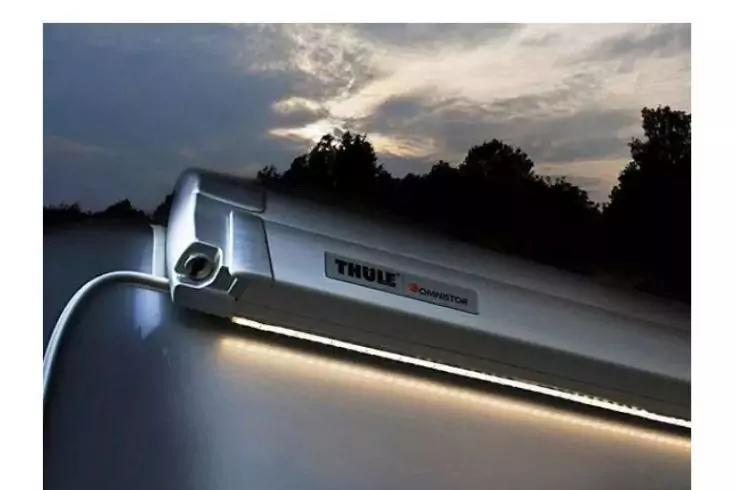 Silbernes Gehäuse mit Thule-Logo, unterseitige LED-Lichtleiste leuchtet, ein Kabel, Abendhimmel mit Wolken und Baum-Silhouetten