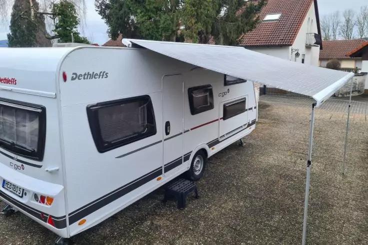 Weißer Wohnwagen (Marke Dethleffs) mit ausgefahrener Markise auf Kiesplatz, Stützstangen, zwei Seitenfenster, Häuser und Bäume im Hintergrund