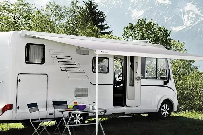 Weißes Wohnmobil mit ausgefahrener Markise, Klapptisch und -stühlen davor, Becher auf dem Tisch, geöffnete Tür, Bäume und Berge im Hintergrund