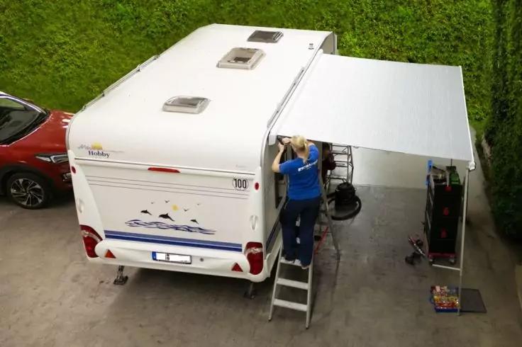Campingwagen mit ausgefahrener Markise, eine Person auf einer Leiter arbeitet daran, im Hintergrund Hecke und ein rotes Auto