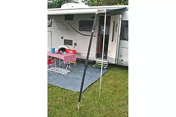 Wohnmobil mit offener Tür, zwei Markisenstützen, graue Bodenmatte, Tisch mit rot-weiß kariertem Tuch, zwei weiße Klappstühle, kleiner roter Kindersitz, grüner Rasen