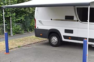 Seiten- und Heckansicht eines weißen Wohnmobils mit ausgefahrener grauer Markise, zwei weißen Stützen und zwei blauen Pfosten auf Parkplatz; im Hintergrund Hecke und Drahtzaun