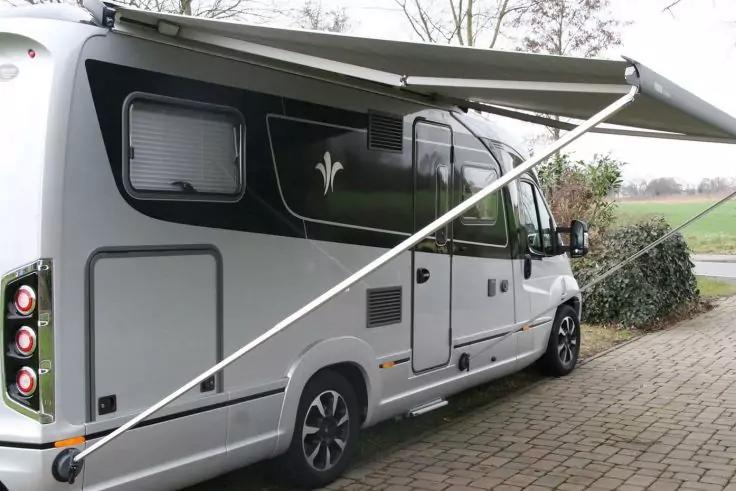 Weißes Wohnmobil von der linken Rückseite, ausgefahrene Markise, Fenster, Seitentür, gepflasterte Fläche, Strauch und Feld im Hintergrund