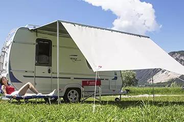 Wohnwagen mit ausgefahrener weißer Markise auf einer Wiese; Person liegt auf einem Liegestuhl daneben, rote Kühlbox vor dem Wohnwagen, Berge und blauer Himmel mit Wolken im Hintergrund