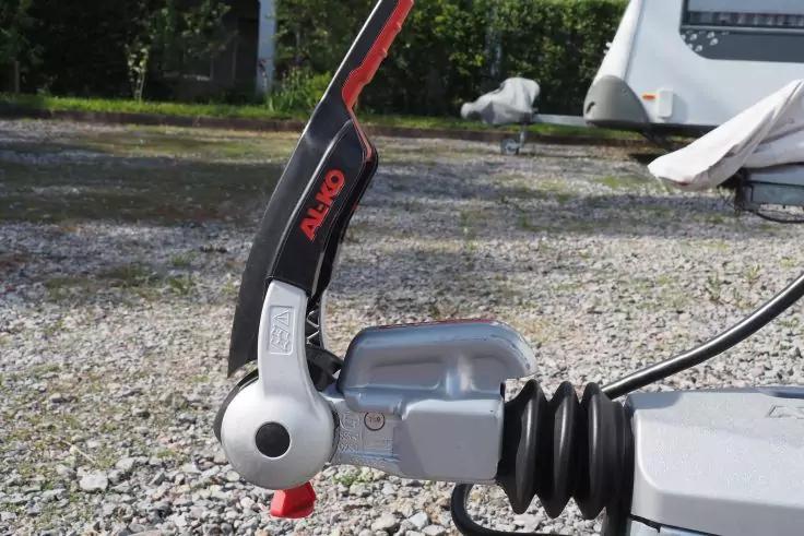 Nahaufnahme einer silbernen Anh&auml;ngerkupplung mit schwarzem Hebel, rotem Knopf und schwarzem Faltenbalg auf kiesigem Untergrund; im Hintergrund ein wei&szlig;er Wohnwagen
