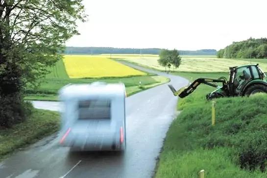Landstra&szlig;e mit unscharf fahrendem Wohnmobil, rechts ein Traktor mit Vorderlader, gr&uuml;ne Wiesen und gelbes Rapsfeld, B&auml;ume am Stra&szlig;enrand
