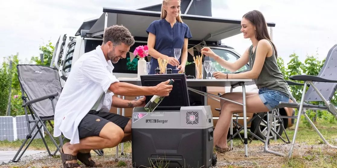 Drei Personen an einem Campingtisch neben einem geparkten Van mit ausgeklapptem Dach; ein Mann kniet und stellt ein rechteckiges tragbares Ger&auml;t mit Display auf den Boden, zwei Frauen sitzen am Tisch, eine gie&szlig;t ein Getr&auml;nk in ein Glas