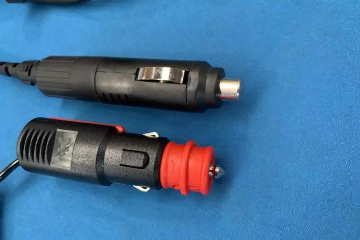 Zwei schwarze Zigarettenanzünder-Stecker, einer mit rotem Kunststoffteil, auf blauem Stoffhintergrund