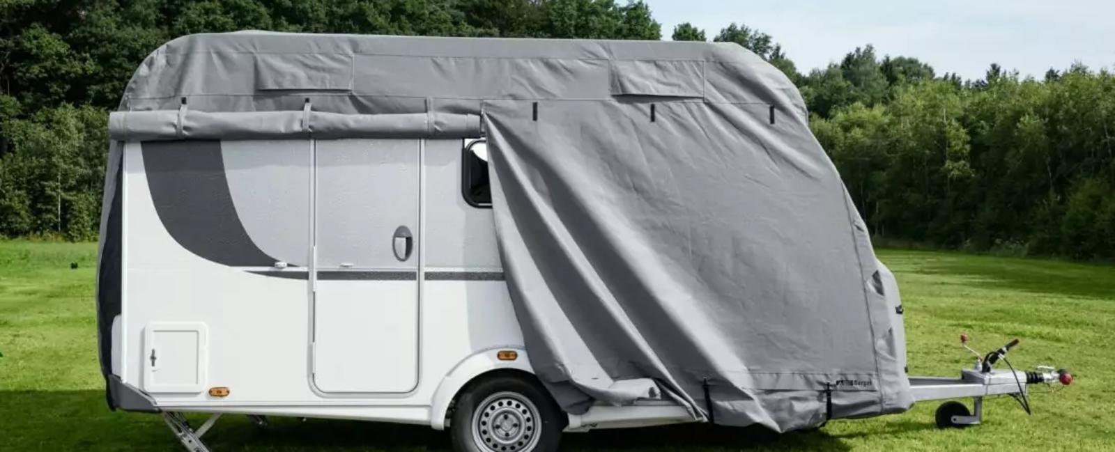 Caravan bianco su un prato, la parte anteriore e parte del fianco coperti da un telo grigio; ruota, porta e gancio di traino visibili; alberi sullo sfondo