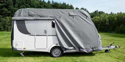 Wei&szlig;er Wohnwagen auf einer Wiese, Vorderteil und teils Seite mit grauer Plane bedeckt; Rad, T&uuml;r und Anh&auml;ngerkupplung sichtbar; Wald im Hintergrund