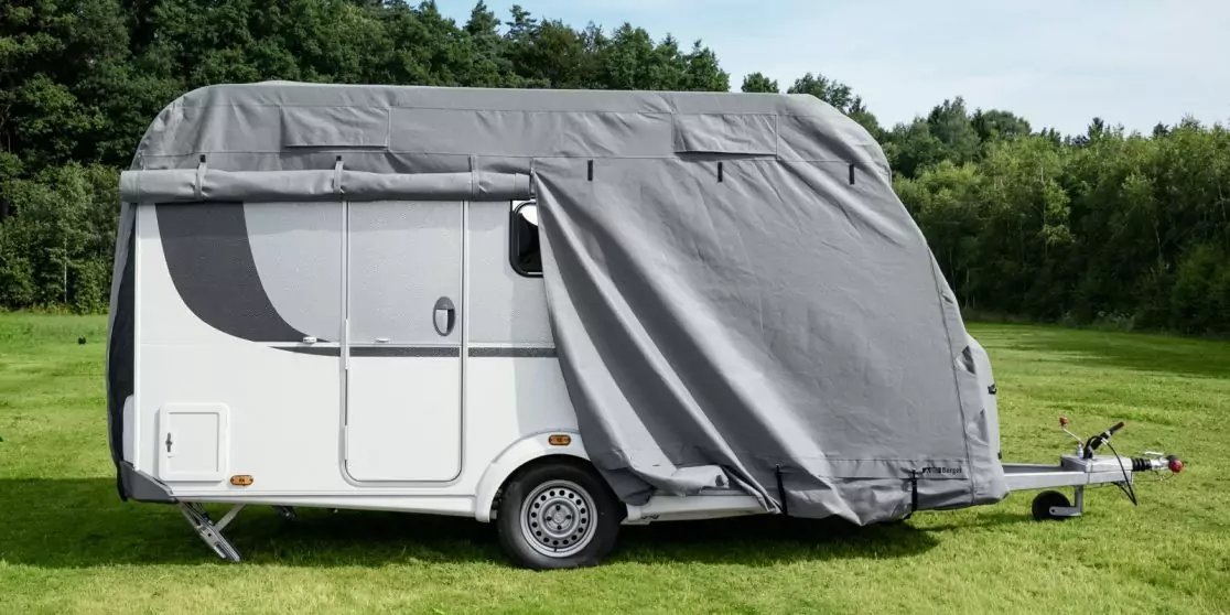 Wei&szlig;er Wohnwagen auf einer Wiese, Vorderteil und teils Seite mit grauer Plane bedeckt; Rad, T&uuml;r und Anh&auml;ngerkupplung sichtbar; Wald im Hintergrund