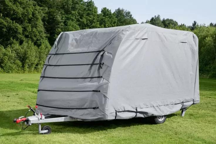 Kleiner einachsiger Anhänger vollständig mit grauer Abdeckung bedeckt, auf einer Wiese vor Bäumen