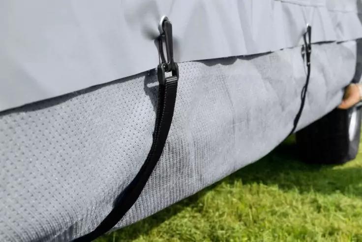 Nahaufnahme einer grauen Abdeckung mit schwarzen Spannbändern und Metallhaken, strukturierter unterer Stoff, grüner Rasen im Vordergrund, rechts teilweise ein Rad und eine Hand
