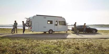 Wei&szlig;es Wohnmobil mit angebundenem Auto auf einer K&uuml;stenstra&szlig;e, mehrere Personen und Fahrr&auml;der sichtbar, Meer im Hintergrund