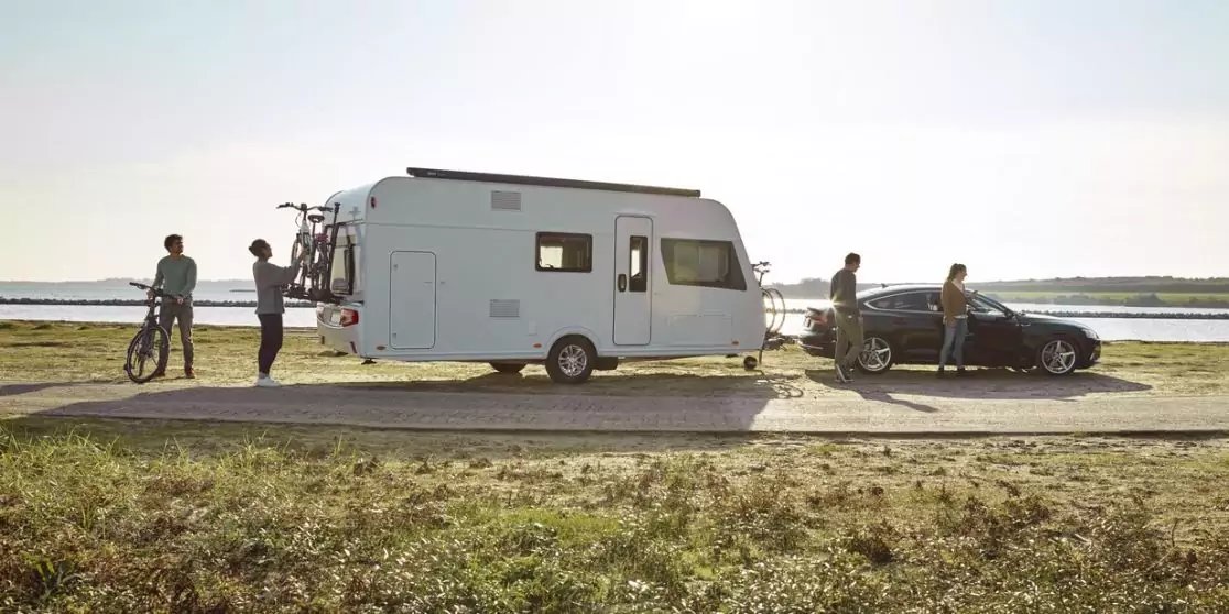 Wei&szlig;es Wohnmobil mit angebundenem Auto auf einer K&uuml;stenstra&szlig;e, mehrere Personen und Fahrr&auml;der sichtbar, Meer im Hintergrund