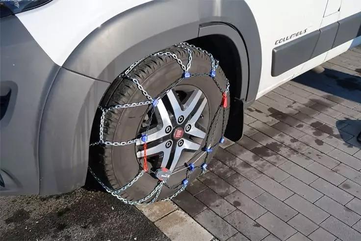 Vorderes Rad eines wei&szlig;en Fahrzeugs mit Metall-Schneeketten, geparkt auf gepflastertem Untergrund