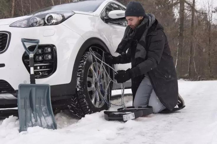 Mann kniet im Schnee neben wei&szlig;em Auto und befestigt Schneekette am Vorderrad; Schneeschaufel und Werkzeugkasten daneben