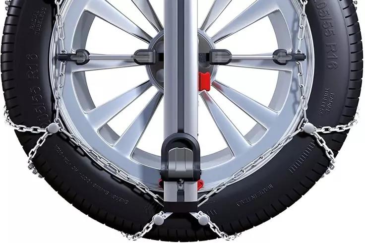 Nahaufnahme eines Autoreifens mit silberner Felge und metallischen Ketten, davor ein senkrechter Metallb&uuml;gel mit rotem Kunststoffteil, wei&szlig;er Hintergrund
