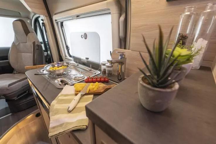 Wohnmobil-K&uuml;chenzeile mit Sp&uuml;le, Zweiflamm-Herd, Schneidebrett mit geschnittener gelber Paprika und Cocktailtomaten, Messer, gestreiftes Tuch, kleine Sukkulentenpflanze, Holzschr&auml;nke und Fenster