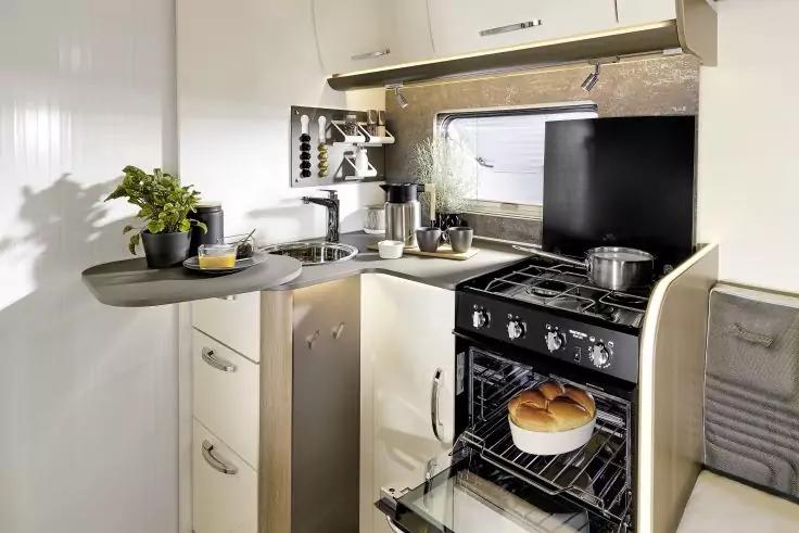 K&uuml;chenzeile mit Sp&uuml;le und Wasserhahn, Arbeitsfl&auml;che mit Topfpflanze und K&uuml;chenutensilien, Kochfeld mit T&ouml;pfen, offener Backofen mit Brot, Fenster dar&uuml;ber