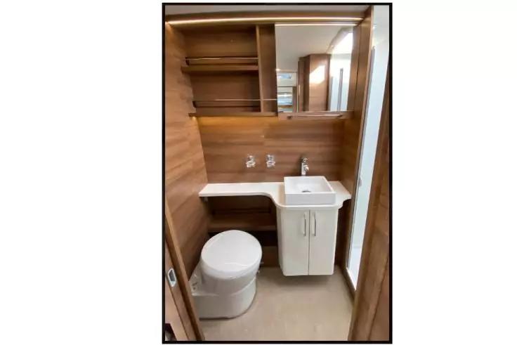 Kleines Badezimmer mit rundem wei&szlig;em WC links, kleinem wei&szlig;em Waschbecken mit Unterschrank rechts, holzvert&auml;felten W&auml;nden, Spiegelschrank mit offenen Regalf&auml;chern dar&uuml;ber, zwei Wandarmaturen &uuml;ber dem Waschbecken und hellem Boden