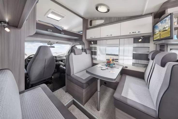 Innenraum eines Wohnmobils mit Fahrer- und Beifahrersitz, Essbereich mit Tisch und gepolsterten Sitzb&auml;nken, Oberschr&auml;nken, Fenster mit Vorhang, an der Wand montiertem Fernseher und Deckenleuchten