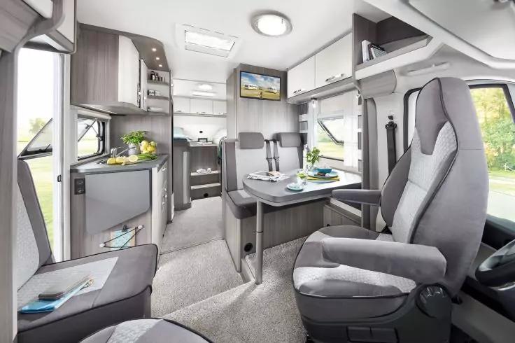 Innenraum eines Wohnmobils mit drehbaren Vordersitzen, einem Tisch mit Besteck und Gl&auml;sern, K&uuml;chenzeile mit Sp&uuml;le und K&uuml;hlschrank, an der Wand montiertem Fernseher, Deckenspots und Fenstern mit Blick auf Gr&uuml;n