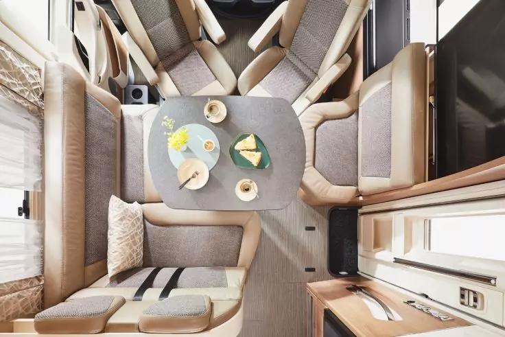 Vogelperspektive auf Innenraum mit mittigem Tisch, gepolsterte Sitzb&auml;nke und St&uuml;hle, Teller, Tassen und Serviette auf dem Tisch, Kissen auf einer Sitzbank, Fenster links, Schr&auml;nke und Arbeitsfl&auml;che rechts