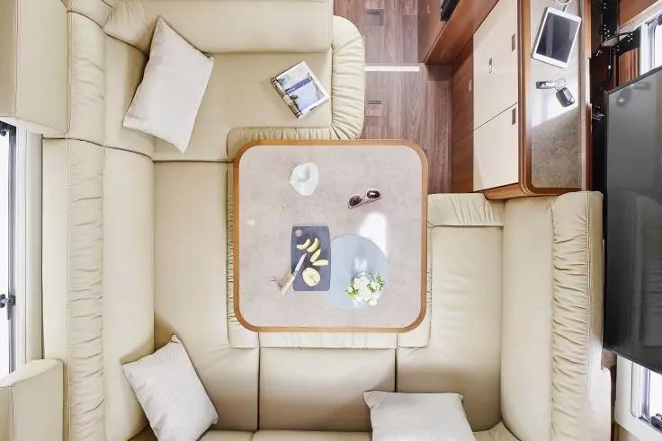 Draufsicht auf eine helle Sitzecke mit beigen Polstersofas um einen quadratischen Tisch. Auf den Sofas wei&szlig;e Kissen und ein flaches rechteckiges Objekt. Auf dem Tisch eine wei&szlig;e Tasse auf Untertasse, ein dunkles rechteckiges Objekt mit gelben Details