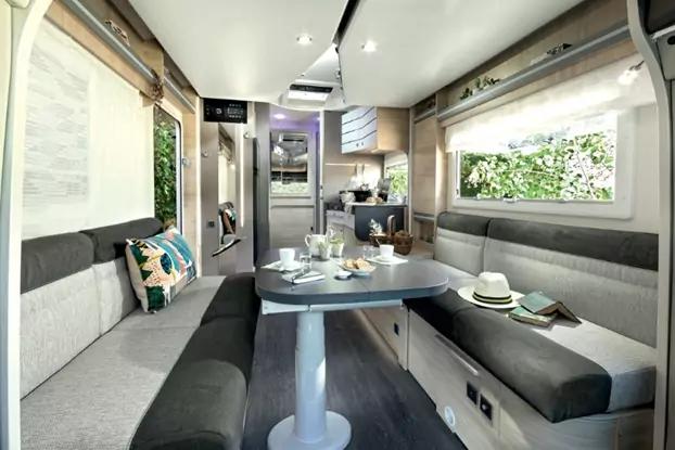 Innenraum eines Wohnmobils mit zwei gegen&uuml;berliegenden Polstersitzb&auml;nken, mittigem Tisch mit Geschirr, K&uuml;chenzeile hinten, Oberschr&auml;nken und Fenstern mit Gr&uuml;n au&szlig;en