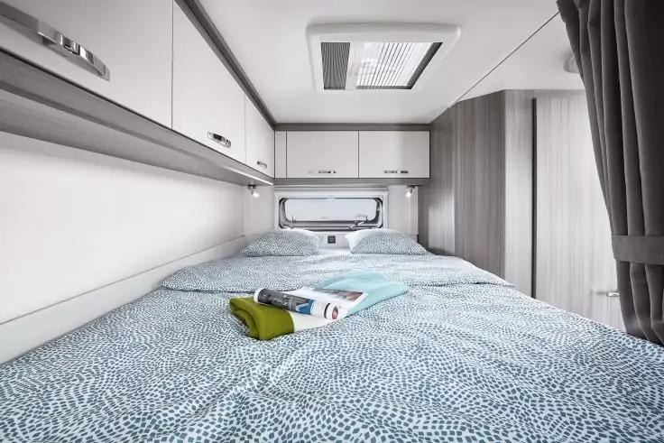 Schlafbereich mit Doppelbett, gemustertem Bettbezug, zwei auf dem Bett liegenden Handt&uuml;chern und einer Zeitschrift; Oberschr&auml;nke, Deckenluke, Fenster, Vorhang und Kleiderschrank