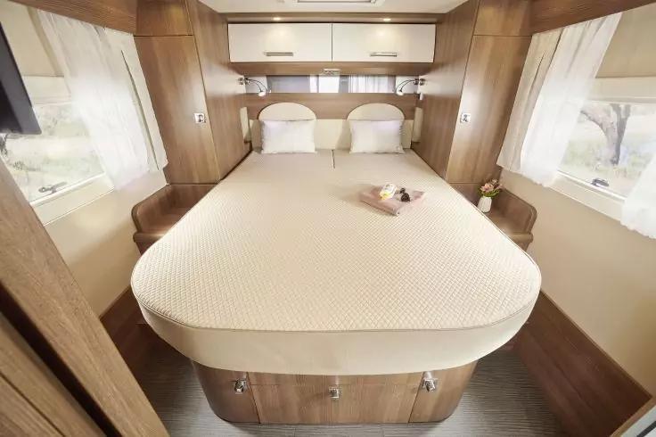 Doppelbett in einem Wohnmobil mit wei&szlig;er Bettdecke, zwei Kopfkissen, Holzschr&auml;nken &uuml;ber dem Kopfende, Fenstern mit Vorh&auml;ngen auf beiden Seiten und einem kleinen Tablett mit Blumen auf dem Bett