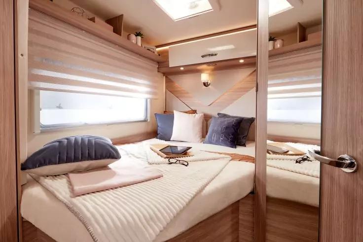 Kompakter Schlafbereich mit Doppelbett, Holzverkleidung, zwei rechteckigen Fenstern mit Rollos, drei Kissen (zwei dunkel, eines hell), Tablet und Brille auf der Bettdecke, Regalf&auml;cher oberhalb, Spiegel an der Seitenwand