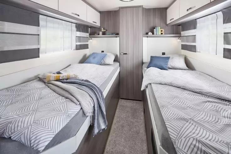 Innenraum mit zwei gegen&uuml;berliegenden Einzelbetten, schmaler Gang dazwischen, Schrankt&uuml;r hinten, Oberf&auml;cher, Fenster mit Vorh&auml;ngen, graue Bettw&auml;sche mit geometrischem Muster, Kissen auf beiden Betten