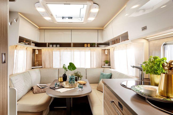 Innenraum eines Wohnmobils mit U-f&ouml;rmiger Polsterbank um einen Tisch; auf dem Tisch Flasche Wein, Gl&auml;ser, Pflanzen und Tablett; Fenster mit Vorh&auml;ngen, Deckenfenster, H&auml;ngeschr&auml;nke; gegen&uuml;ber K&uuml;chenzeile mit Sp&uuml;le, Arbeitsfl&auml;che und Topf mit Kr&auml;uterpf