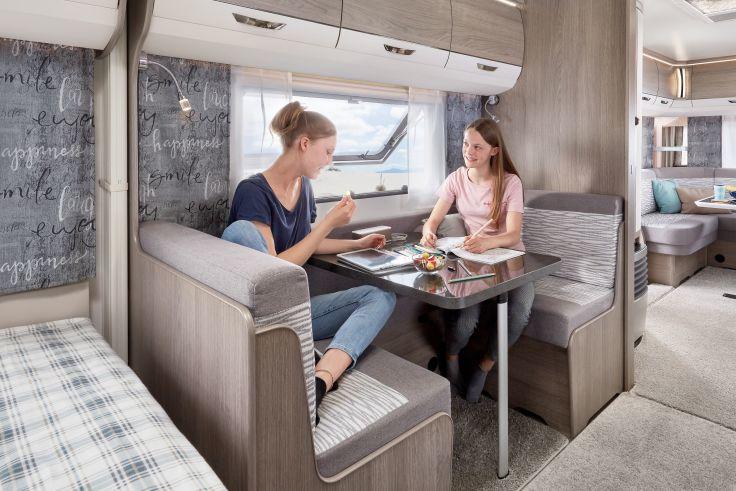 Zwei Personen sitzen an einem Tisch im Innenraum eines Wohnmobils; auf dem Tisch Teller und eine Zeitschrift, Sitzb&auml;nke, Fenster, sichtbarer Schlaf- und Kochbereich