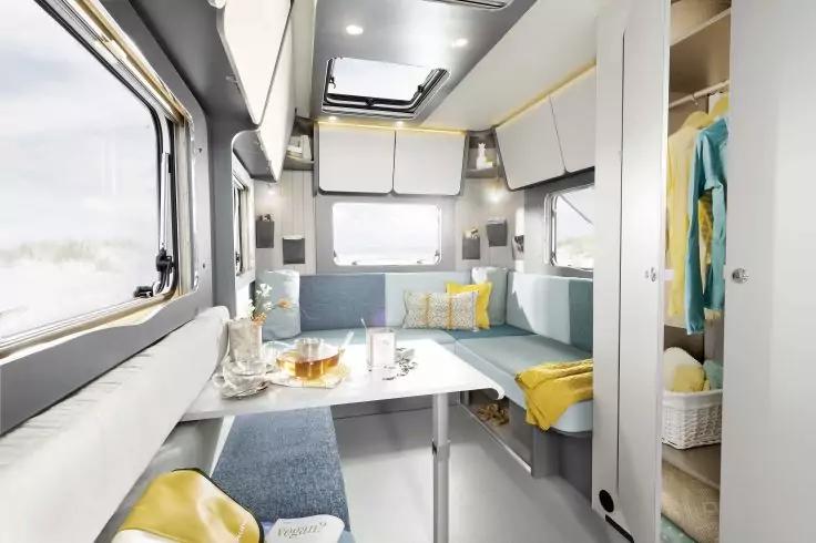 Innenraum eines Wohnmobils mit Essbereich: Tisch mit Gl&auml;sern und Teekanne, umliegende Sitzb&auml;nke mit Kissen und gelber Decke, Fenster, Ober- und Seitenschr&auml;nke, Deckenluke, offener Kleiderschrank mit h&auml;ngender Kleidung und Korb