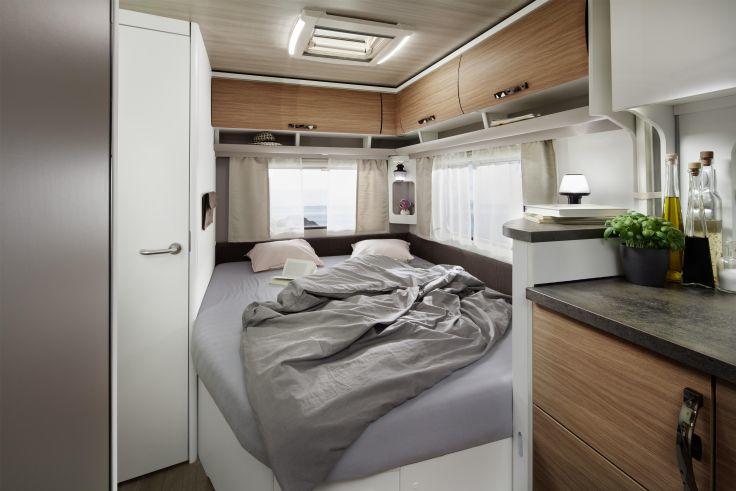 Innenraum eines Wohnmobils: Bett mit zerw&uuml;hlter Decke und Kopfkissen, seitliche Fenster mit Vorh&auml;ngen, Holzschr&auml;nke, kleine Arbeitsfl&auml;che mit Pflanze und Flaschen, Deckenluke