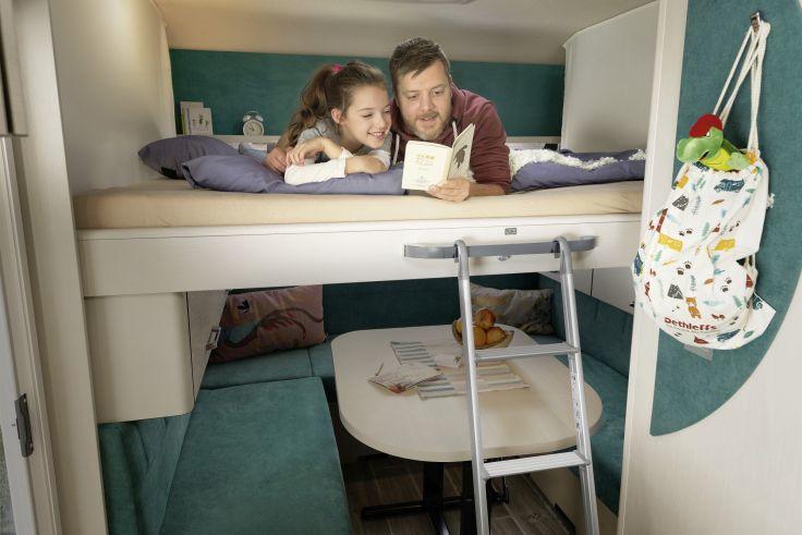 Ein Mann und ein Kind liegen auf dem oberen Etagenbett und lesen ein Buch. Unter dem Bett sind ein Tisch mit Gegenst&auml;nden, eine Leiter und ein h&auml;ngender Rucksack sichtbar