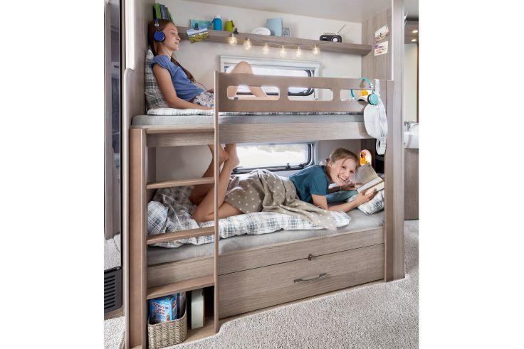 Etagenbett aus hellem Holz in kompakter Schlafnische; oben ein Kind mit Kopfh&ouml;rern, unten ein Kind mit Tablet; Leiter, Regale, Fenster