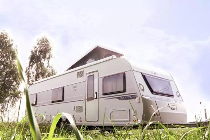 Wei&szlig;er Wohnwagen auf einer Graswiese, B&auml;ume im Hintergrund, heller Himmel mit vereinzelten Wolken