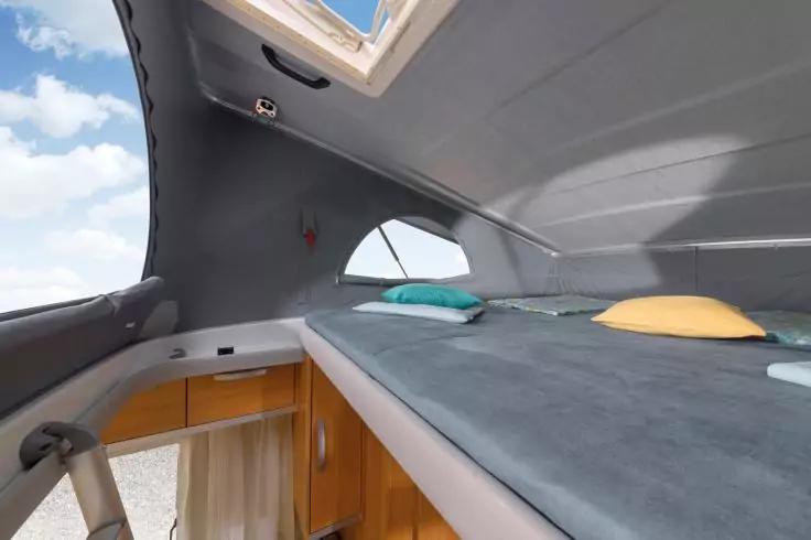 Erh&ouml;hte Liegefl&auml;che mit grauem Matratzenbezug, zwei Kissen (t&uuml;rkis und gelb), ge&ouml;ffnetes Dachfenster mit Blick auf den Himmel, kleines seitliches Fenster und Holzschrank darunter