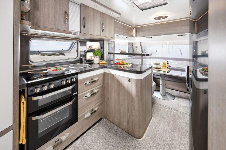 Innenraum einer Wohnwagenk&uuml;che mit Herd und Backofen, Sp&uuml;le, Arbeitsfl&auml;chen, H&auml;ngeschr&auml;nken, Fenster, Esstisch mit Polsterbank, Obstschale und Blumenvasen