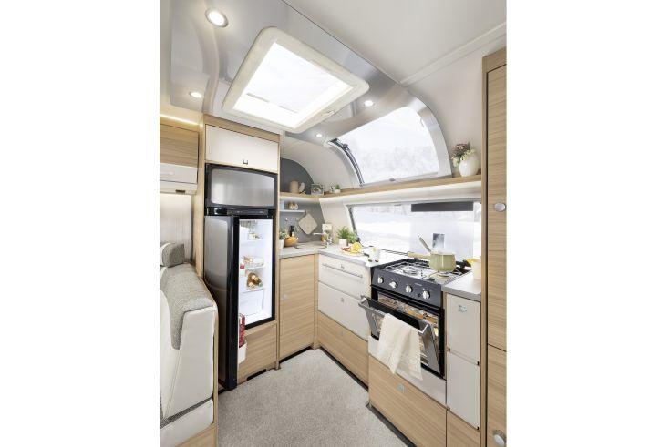 Innenraum mit kompakter K&uuml;chenzeile: Herd mit Ofen und Handtuch, Sp&uuml;le, ge&ouml;ffneter K&uuml;hlschrank, helle Holzschr&auml;nke, Dachfenster, Sitzbank und Teppich