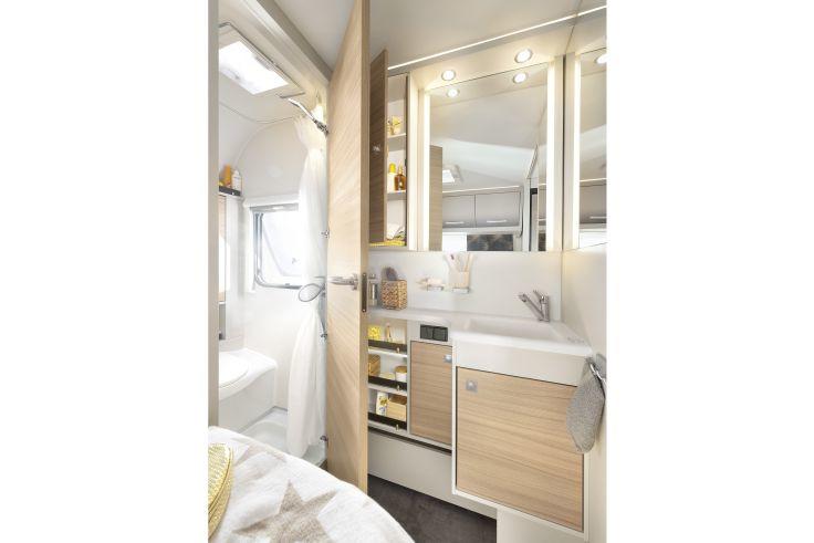 Kleines Badezimmer mit Waschbecken, Unterschrank und beleuchtetem Spiegel, Duschbereich, Toilette, Regalf&auml;cher mit Pflegeprodukten und h&ouml;lzernen Schrankfronten