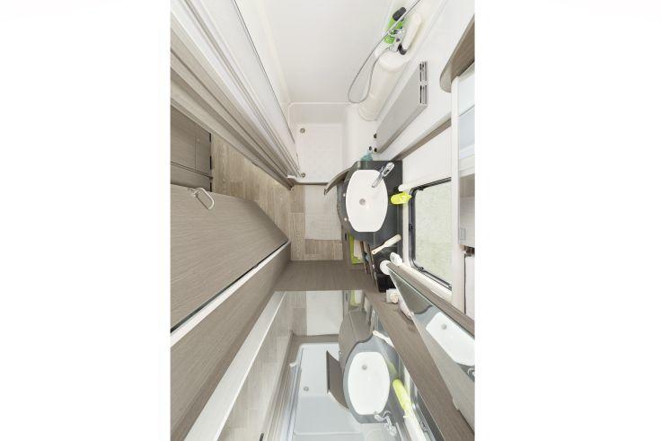 Wei&szlig;es ovales Waschbecken mit Chromarmatur, runder Spiegel, dunkle gl&auml;nzende Arbeitsfl&auml;che, Handbrause mit Schlauch, wei&szlig;er Duschvorhang, graue Wandfliesen, gr&uuml;ner Seifenspender, wei&szlig;e T&uuml;r